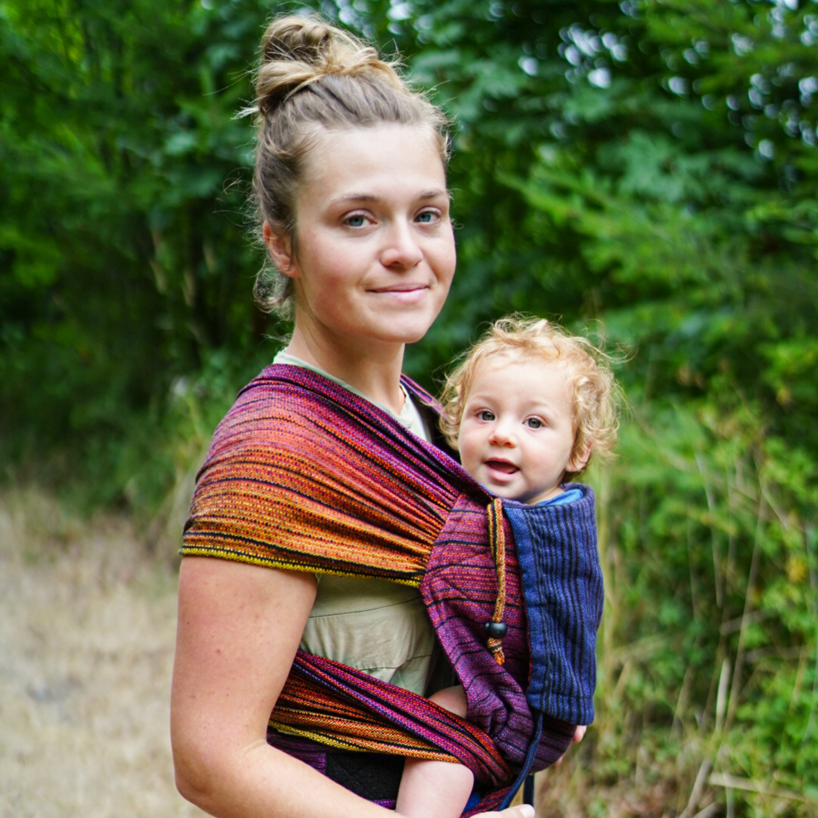 Baby in Trage, getragen von der Mama. WrapMySol Twilight. Im Hintergrund grüne Büsche